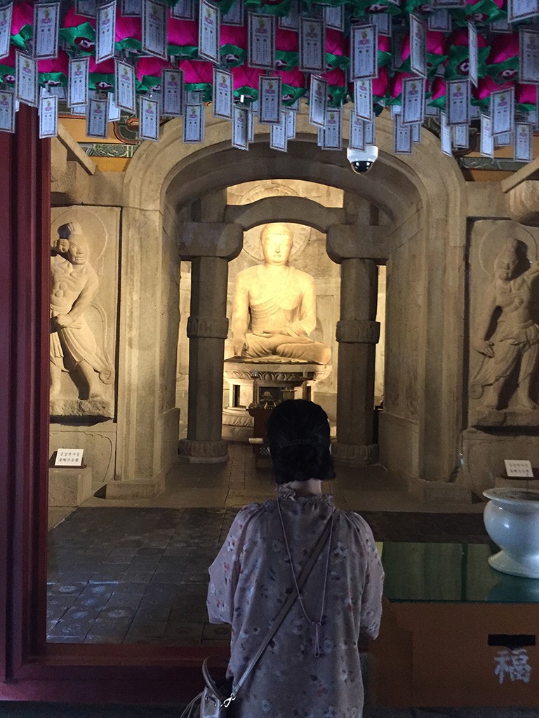 A woman in a floral-patterned dress stands in quiet prayer before the serene Buddha statue inside Seokguram Grotto, surrounded by ancient stone carvings and glowing prayer lanterns.